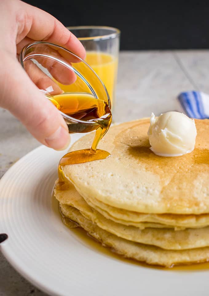 stack of homemade pancakes with butter on a plate being covered with maple syrup