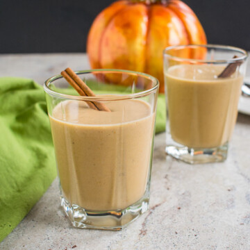 two glasses of pumpkin smoothies with cinnamon sticks in front of a pumpkin and napkins