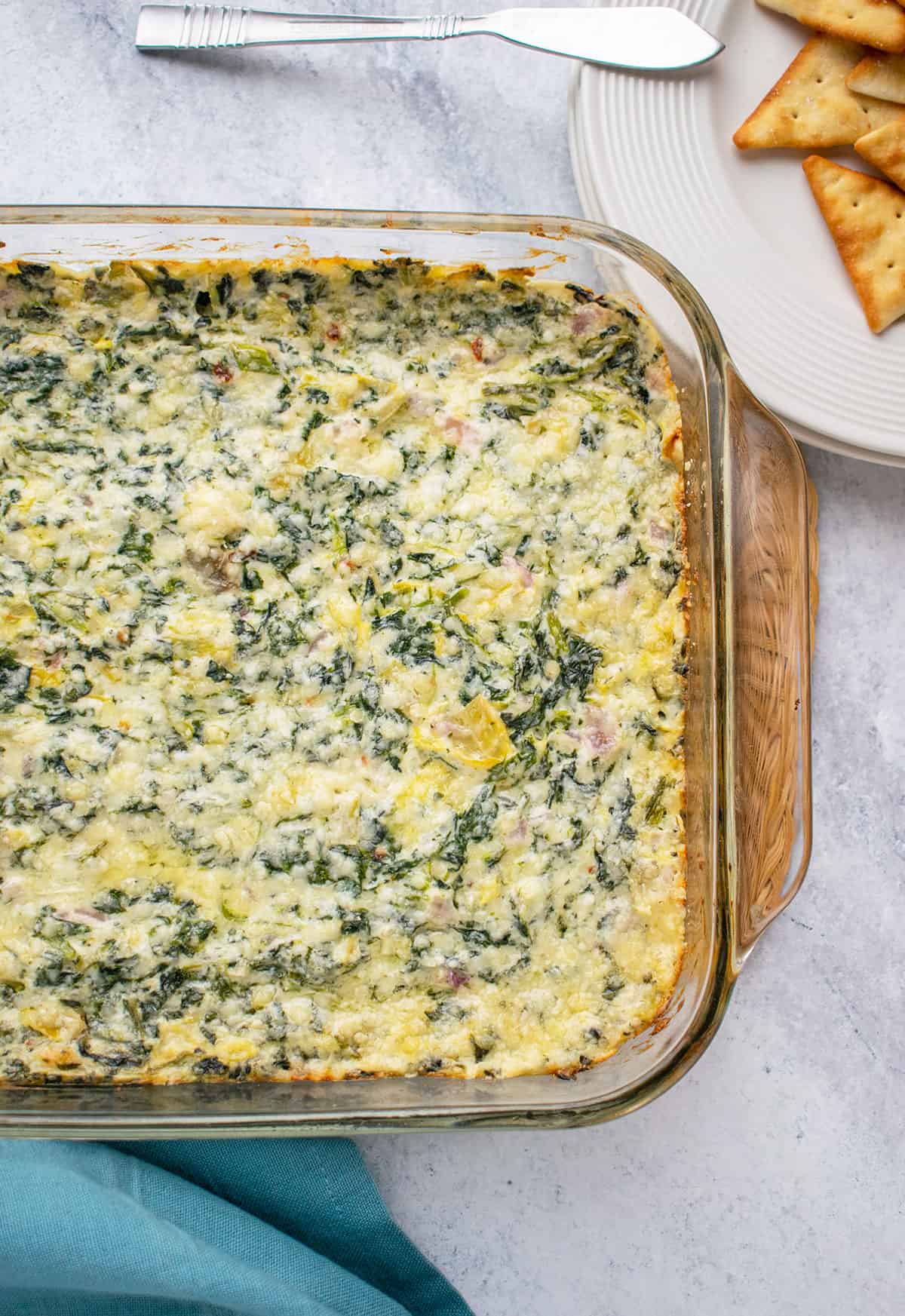 Overhead photo of Asiago Spinach and Artichoke Dip in pan with plate of pita crackers