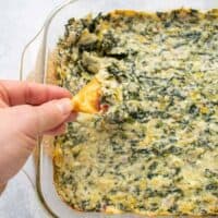 Overhead photo of hand holding a pita cracker topped with Asiago Spinach and Artichoke Dip over the baking dish