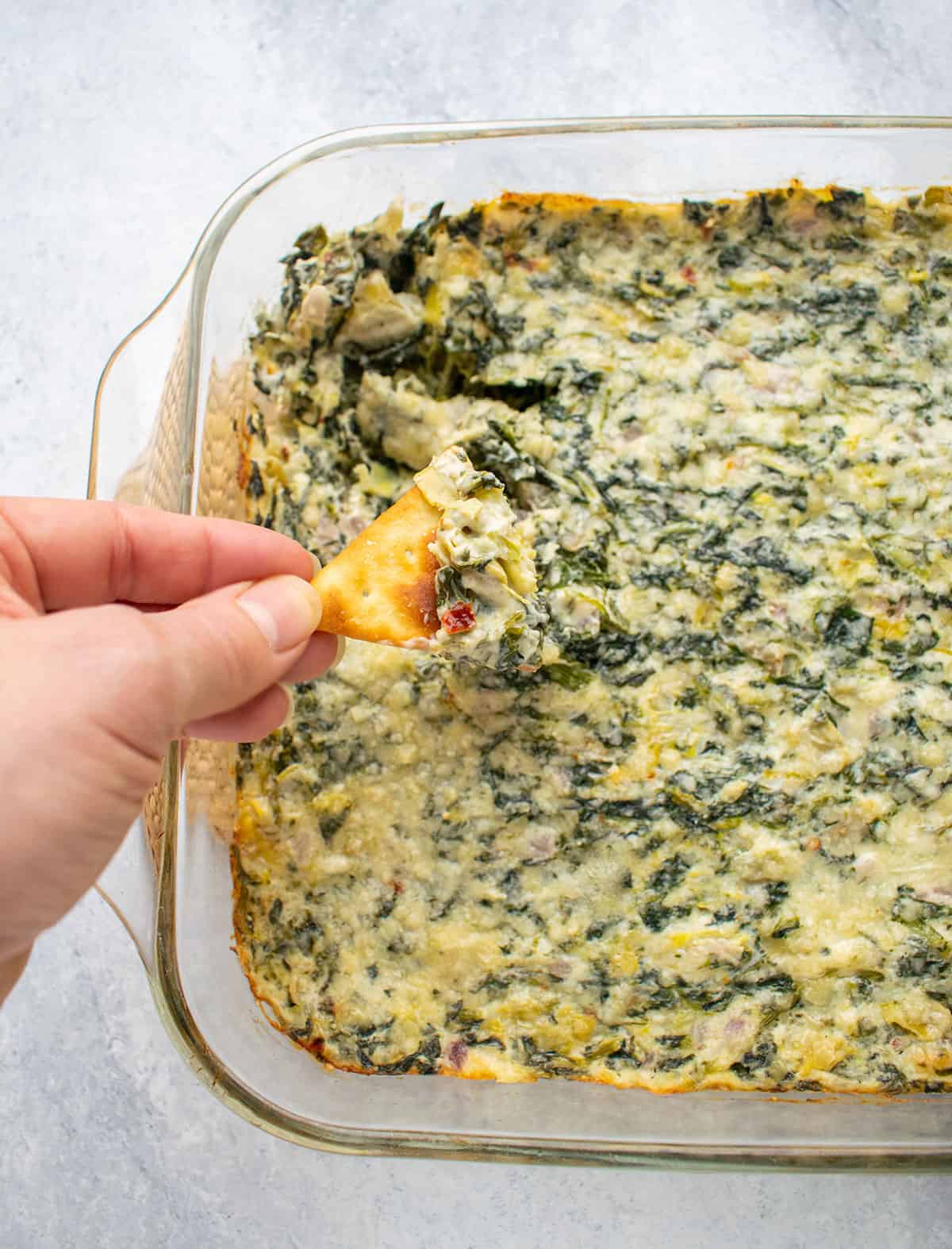 Overhead photo of hand holding a pita cracker topped with Asiago Spinach and Artichoke Dip over the baking dish