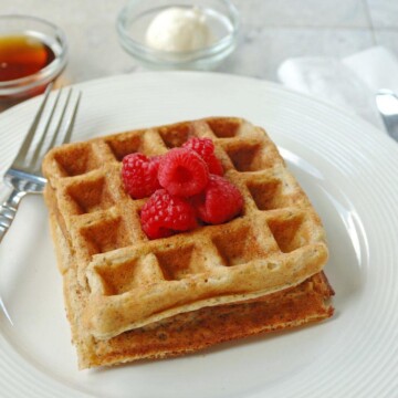 2 Buttermilk Whole Grain Waffles stacked on a plate with raspberries, fork, butter and syrup