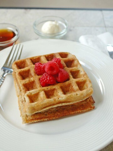 2 Buttermilk Whole Grain Waffles stacked on a plate with raspberries, fork, butter and syrup