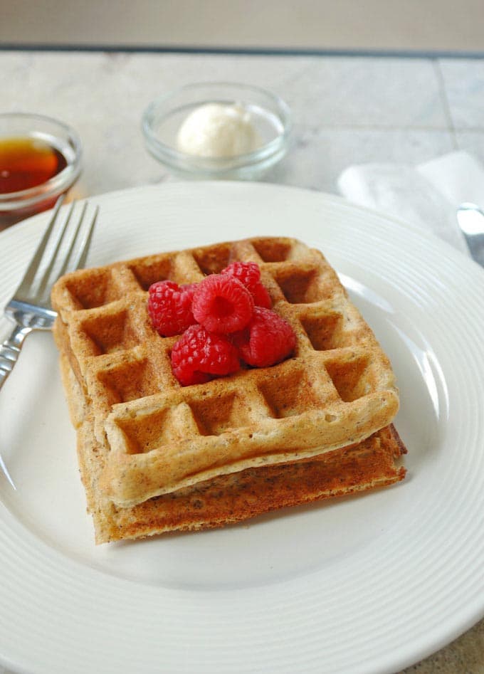 2 Buttermilk Whole Grain Waffles stacked on a plate with raspberries, fork, butter and syrup