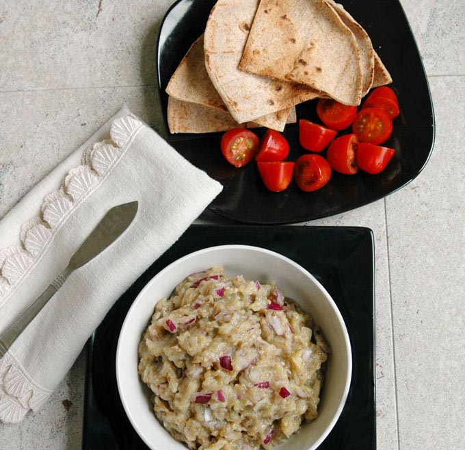 overhead view of Roasted Eggplant Dip in a bowl next to a plate with pita and small tomatoes, napkin with spreading knife