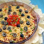 7-Layer Taco Dip in a glass container surrounded by tortilla chips