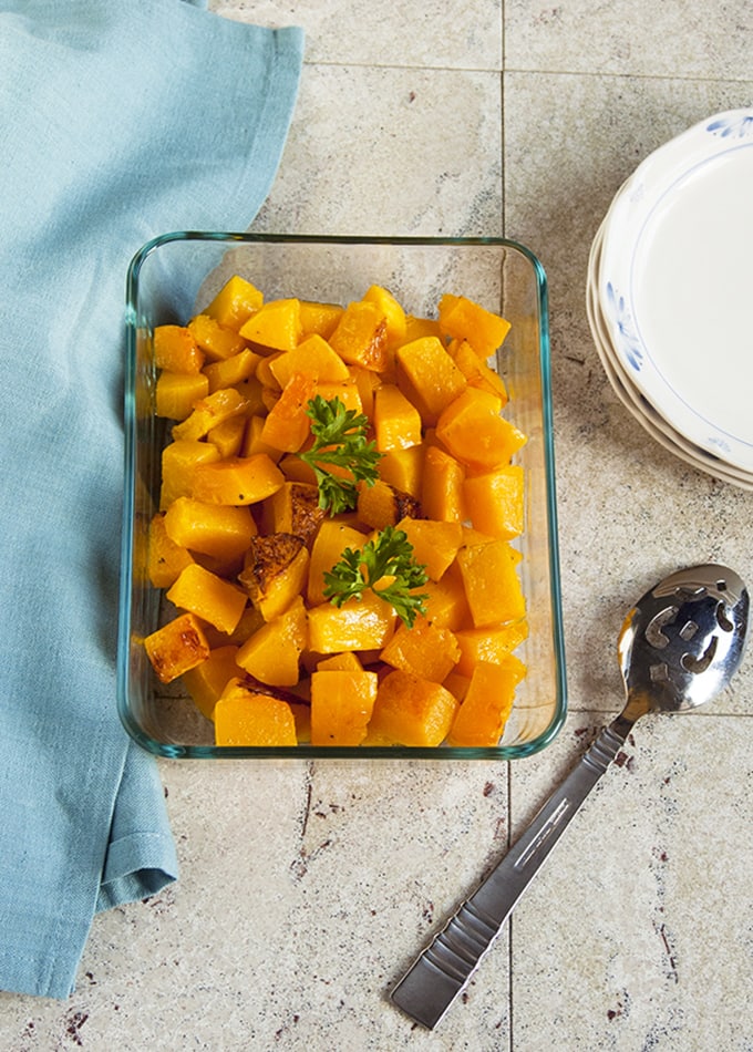 Roasted Butternut Squash Cubes in a glass pan beside serving spoon, plates and napkin