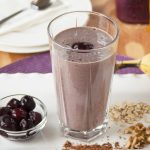 glass of chocolate-covered cherry almond smoothie surrounded by cherries, cocoa, walnuts and oats