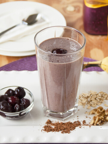 glass of chocolate-covered cherry almond smoothie surrounded by cherries, cocoa, walnuts and oats