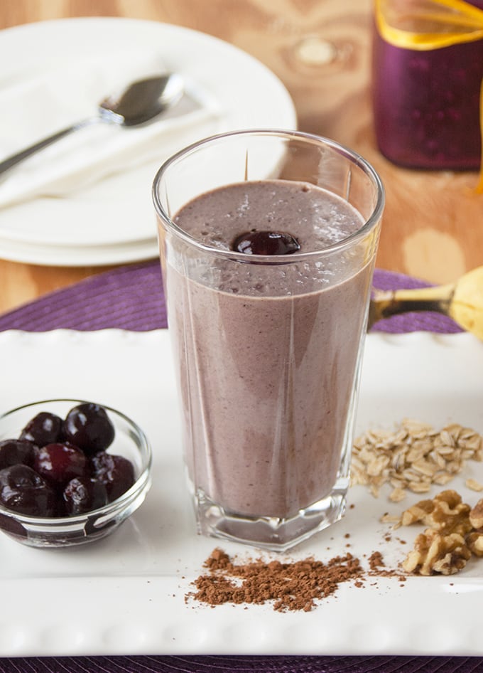 glass of chocolate-covered cherry almond smoothie surrounded by cherries, cocoa, walnuts and oats