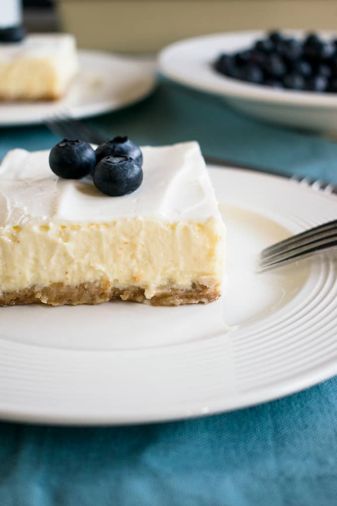 Cheesecake with graham cracker crust and sour cream topping on a plate with three blueberries on top 