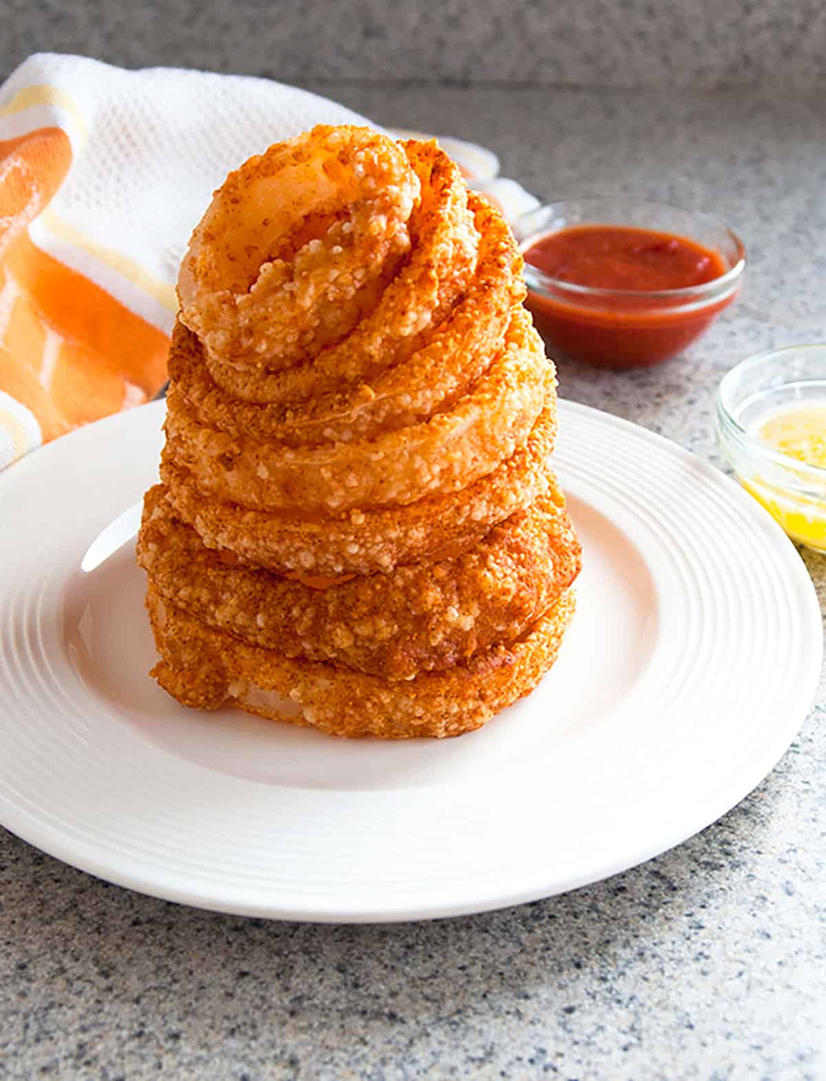 stack of baked parmesan gluten-free onion rings on a plate with dipping sauces behind 