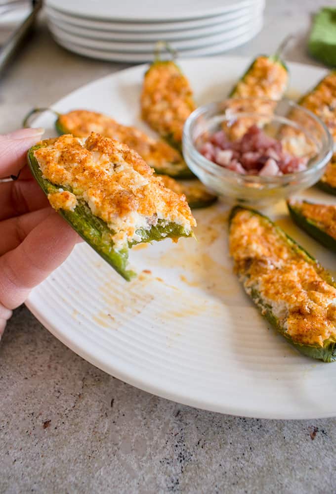 hand holding a baked jalapeno popper with a bite taken out of it in front of a full plate with prosciutto 