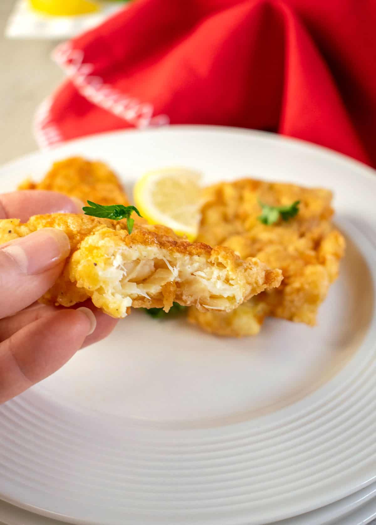 Hand holding bitten piece of fried baccala (salted cod fish) in front of a plate of fried baccala