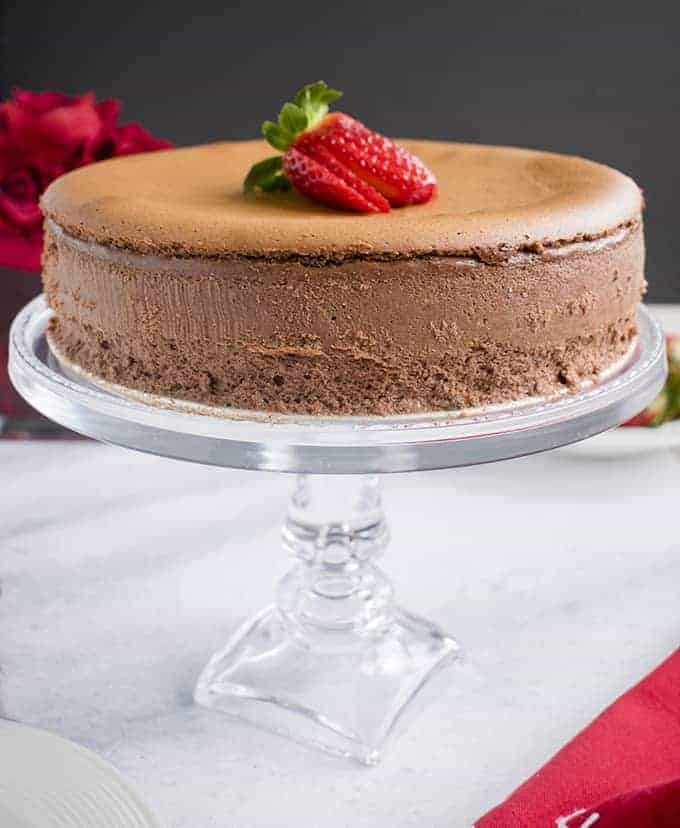 Photo of a gluten free chocolate cheesecake topped with a strawberry on glass cake stand