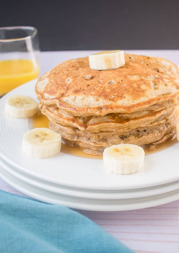 Side view of stacked banana pancakes with banana slices on a plate and a glass of orange juice behind it