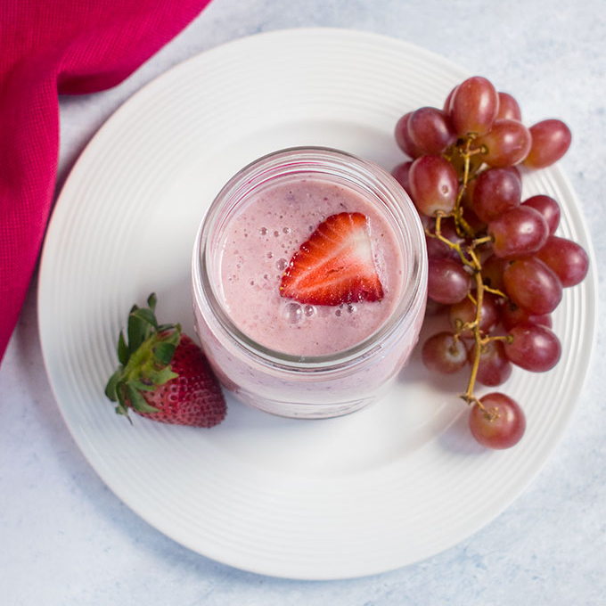 overhead view of pink smoothie with strawberries, grapes, white plate, pink napkin