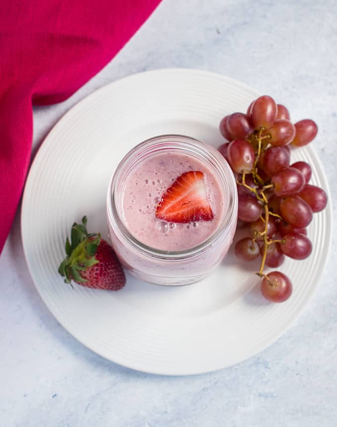 overhead of pink smoothie, grapes, strawberries, white plate, pink napkin