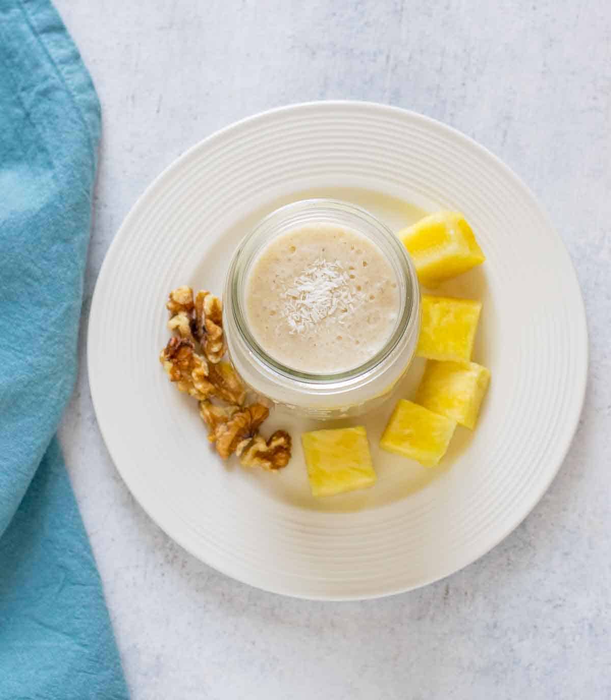 smoothie in a jar with coconut, on plate with pineapple and walnuts