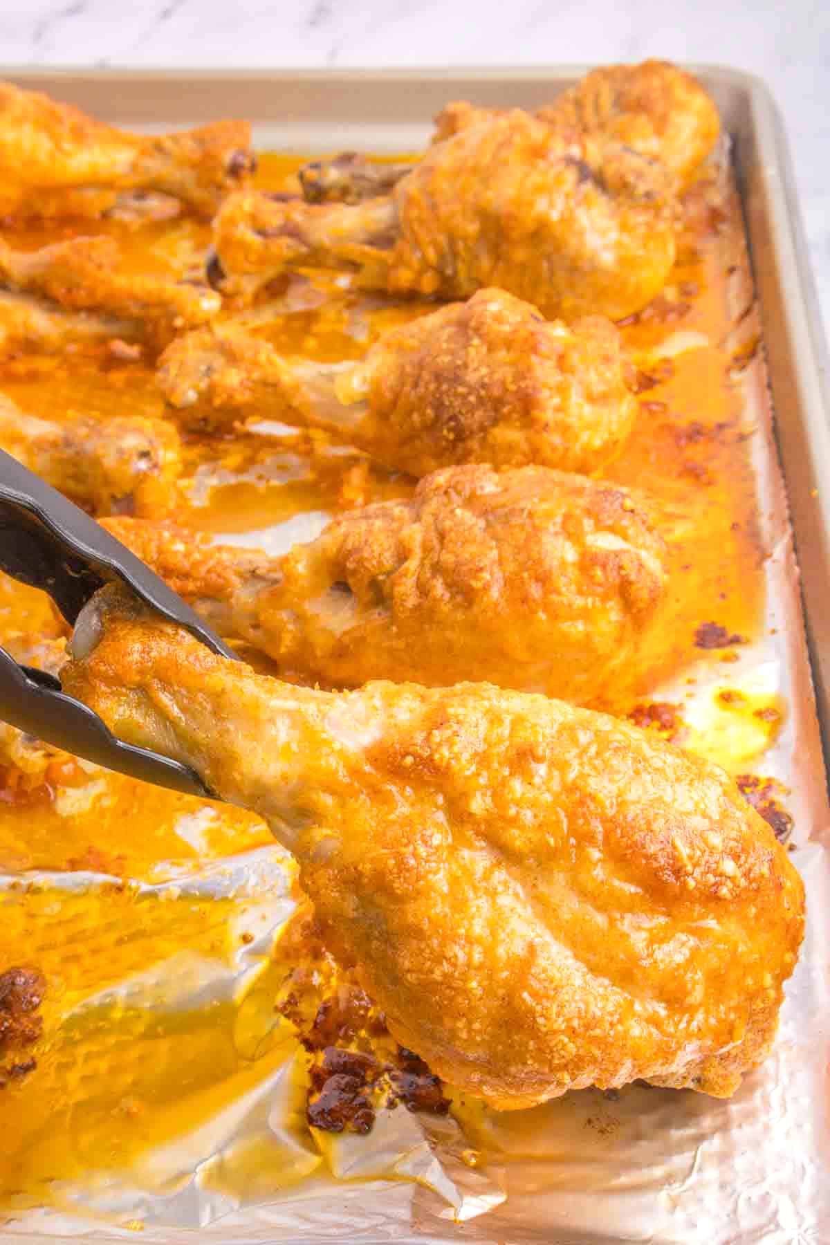 tongs lifting baked chicken drumstick from sheet pan of chicken legs