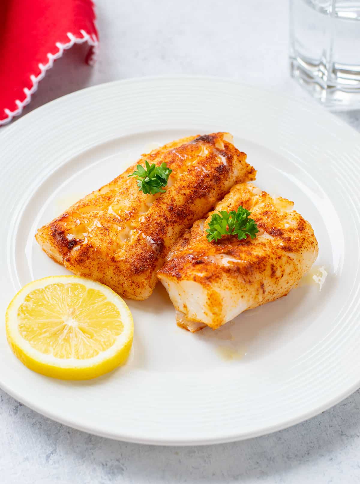 two pieces of grilled, seasoned cod on plate with lemon and parsley.