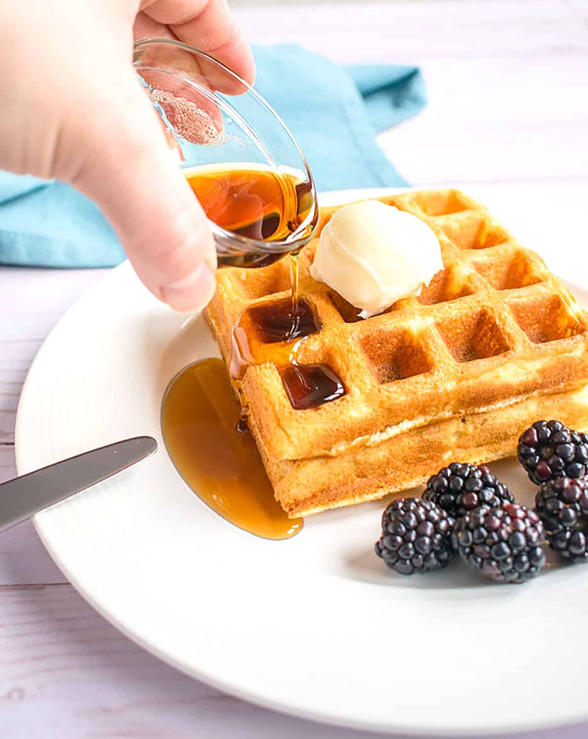 syrup being poured over homemade waffles, butter and blackberries.