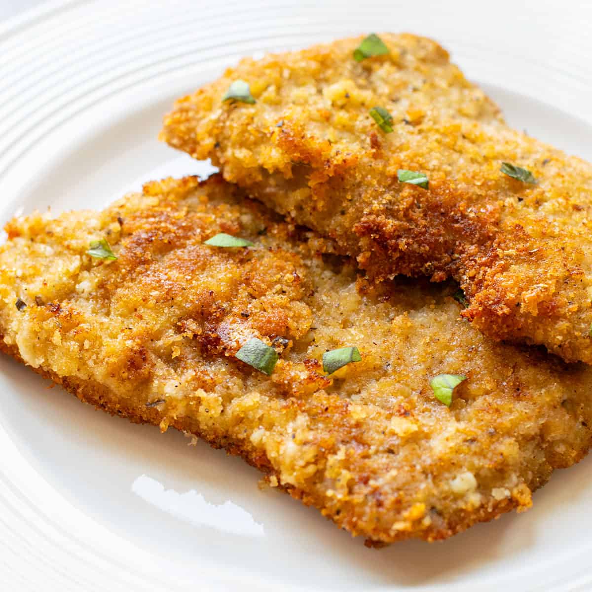 two breaded veal cutlets on a plate topped with basil pieces